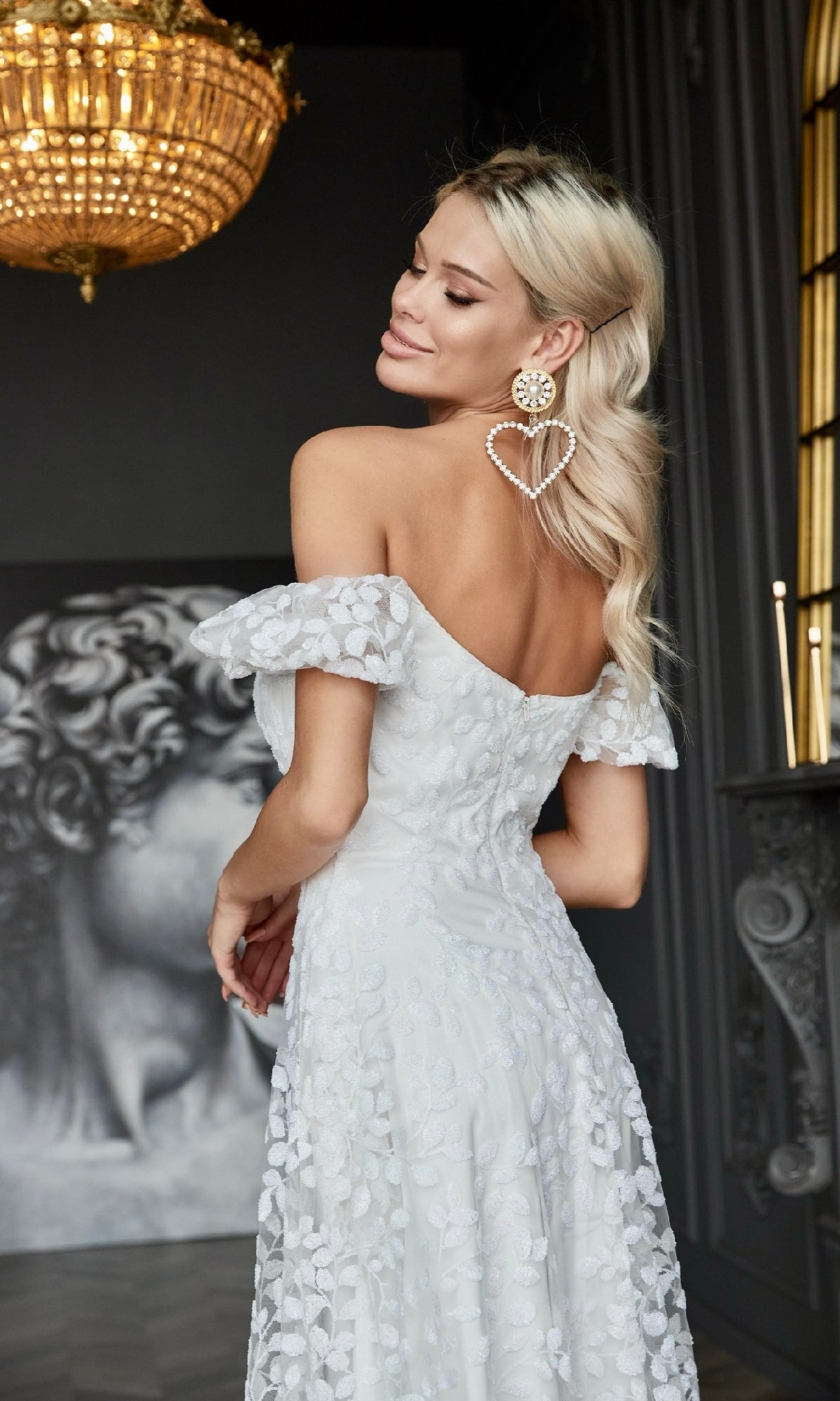 A woman with long blond hair and heart-shaped earrings stands indoors in the Tina Holly TK023W, a long white lace formal dress with floral details, showing her back. A classical statue is seen in the background.